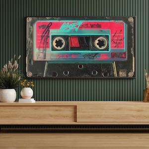 May include: A framed artwork featuring a retro cassette tape design. The cassette has a red and black color scheme with neon blue accents and graffiti-style text. The artwork is mounted on a dark green wall above a wooden cabinet.