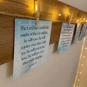 Può includere: Una trave di legno con piccole luci bianche, con diverse carte appese. Le carte sono tenute da mollette di legno e presentano un testo da Sofonia 3:17. Le carte hanno uno sfondo acquerello azzurro chiaro.