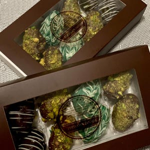 May include: Two boxes of chocolate covered strawberries. The top box has a green and white design with the text "Firdaws Foods" on the label. The bottom box has a green and white design with the text "Firdaws Foods" on the label.