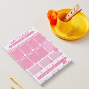 May include: A pink weekly planner notepad with heart motifs and the text "WEEKLY PLANNER" printed at the top. Next to it is a yellow desk organizer containing a wooden ruler showing 6 inches, a pencil, and several metal paper clips.
