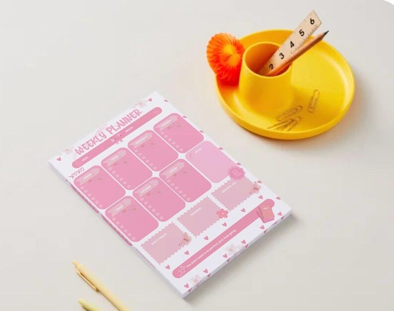May include: A pink weekly planner notepad with heart motifs and the text "WEEKLY PLANNER" printed at the top. Next to it is a yellow desk organizer containing a wooden ruler showing 6 inches, a pencil, and several metal paper clips.