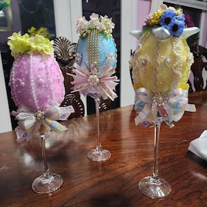 Może przedstawiać: Trzy ozdobne jajka wielkanocne na szklanych nóżkach. Jajka są pokryte brokatem i koralikami, a każde ma inny schemat kolorystyczny: różowy, niebieski i żółty. Jajka są ozdobione kwiatami i wstążkami.