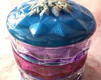 May include: A decorative jar with a blue, purple, and pink color scheme. The lid is blue with a diamond pattern and adorned with seashells. The jar is made of glass and is suitable for storing small items. The jar measures approximately 6 inches tall.