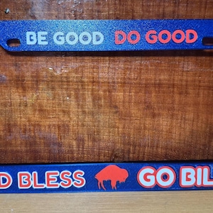 May include: Blue license plate frame with white and red text that reads "BE GOOD DO GOOD" at the top and "GOD BLESS" and "GO BILLS!!" at the bottom. A red buffalo logo is in the center.