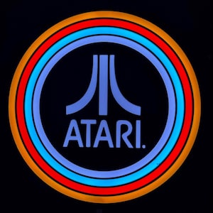 May include: Circular Atari logo with a black background. The logo features a blue Atari symbol inside a blue circle, surrounded by red and orange rings. The word "ATARI" is written in blue below the symbol.