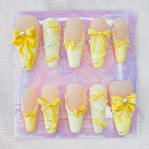May include: A set of ten artificial nails in a variety of designs. Each nail features a yellow base with decorative elements such as bows, pearls, and checkered patterns. The nails are arranged on a marbled, pink and purple surface.