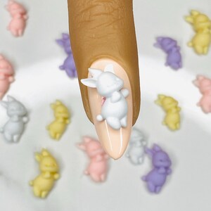 May include: A fingernail with light pink nail polish and a white bunny charm. Surrounding the nail are small, pastel-coloured bunny charms in pink, yellow, grey, and purple, creating an Easter theme.