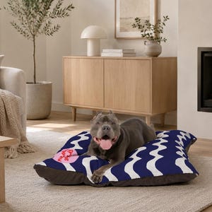 Può includere: Un cane grigio con la lingua di fuori che riposa su una cuccia quadrata per animali. La cuccia presenta una parte superiore in tessuto blu scuro con un motivo ripetuto di linee ondulate bianche e una piccola toppa circolare rosa a sinistra.