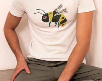 May include: White crew neck t-shirt featuring a stylized bee graphic with yellow and black stripes, and text-based wings. The bee has a pink eye and blue antennae. The model is wearing olive green pants and black sneakers.