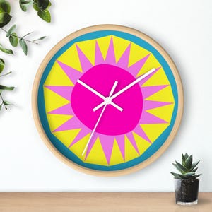 Könnte beinhalten: Eine runde Wanduhr mit Holzrahmen. Das Zifferblatt hat ein lebendiges Design mit einem pinkfarbenen Zentrum, einem gelb-rosa Sternmuster und einem blauen Rand. Weiße Zeiger zeigen die Uhrzeit an.