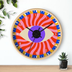 Puede incluir: Un reloj de pared redondo con marco de madera. La esfera del reloj presenta un gran diseño de ojo con iris púrpura, ojo beige y líneas radiantes rojas, rosas y naranjas. Las manecillas negras indican la hora.