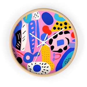 May include: Round wall clock with a light wood frame. The clock face features a pink background with a colorful abstract design of blue, yellow, red, and black shapes. White clock hands indicate the time.