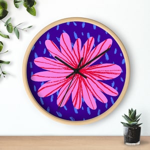 May include: A round wall clock with a light wood frame. The clock face features a large pink flower design with red details against a dark blue background. Black clock hands indicate the time. Decorative home decor.