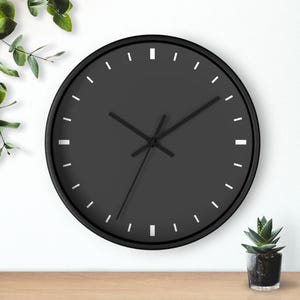 Op de afbeelding: Een ronde, zwarte wandklok met witte, rechthoekige uurmarkeringen en zwarte wijzers. De klok heeft een zwart frame en een donkergrijze wijzerplaat. De klok wordt weergegeven op een houten oppervlak met een witte achtergrond.
