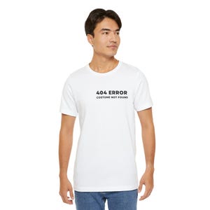 Puede incluir: Camiseta blanca con el texto "404 ERROR COSTUME NOT FOUND" en negro. La camiseta es de manga corta y tiene cuello redondo. El modelo lleva vaqueros azules.