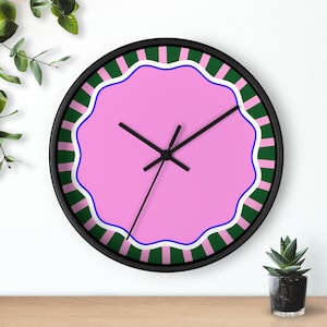 May include: A round wall clock with a black frame. The clock face is pink with a scalloped edge, a blue border, and a green and white striped outer ring. Black clock hands indicate the time. A small potted plant sits below the clock.
