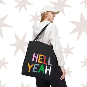 May include: Black tote bag with the words "HELL YEAH" in colorful block letters. The bag has a shoulder strap and is being carried by a person wearing a white hoodie and a baseball cap.
