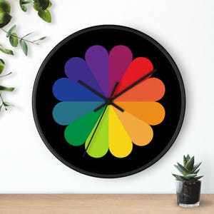 May include: A round wall clock with a black frame and a black face. The clock face features a colorful, petal-like design in rainbow hues. The clock hands are black.