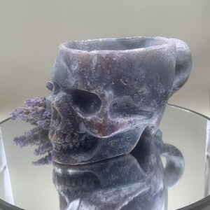 May include: A skull-shaped mug in shades of gray and purple, with a handle on the side. The mug is made of a stone-like material and sits on a reflective surface. A small bundle of dried lavender is placed near the mug.