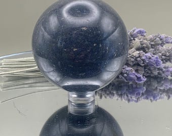 May include: A dark blue crystal ball, resting on a clear stand, reflects on a mirrored surface. The sphere is smooth and polished, with subtle variations in color. A bundle of dried lavender with purple flowers is in the background.
