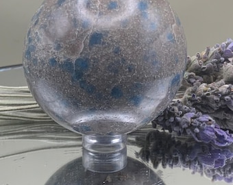 May include: A spherical, decorative object with a speckled pattern of blue and gray hues. The sphere is mounted on a clear, cylindrical stand, reflecting on a mirrored surface. Dried lavender sprigs are visible in the background.
