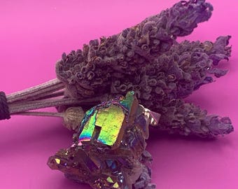 May include: A large iridescent crystal with a rough, faceted surface. The crystal is a deep purple color with hints of green and gold. It is sitting on a pink surface with dried lavender flowers.