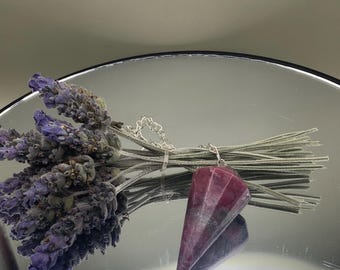 Pode incluir: Um pêndulo de cristal rosa escuro facetado com uma corrente prateada está pendurado ao lado de um feixe de lavanda seca. O pêndulo tem formato de cone e reflete a luz. A lavanda tem flores roxas e caules verdes.