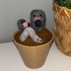 以下が含まれることがあります： 茶色の紙の鉢に入った、手作りのフェルト彫刻。生き物は灰色の体で、ピンクの鼻と黒い目があります。鉢は茶色のフェルトで満たされています。編まれたバスケットと緑の植物が背景にあります。