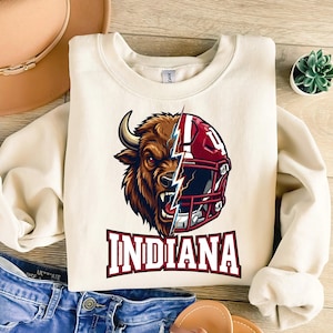 Puede incluir: Sudadera de color crema con un gráfico de una cabeza de bisonte y un casco de fútbol dividido por un rayo. La palabra "INDIANA" está impresa en letras de bloque granate debajo del gráfico. La sudadera tiene mangas largas.