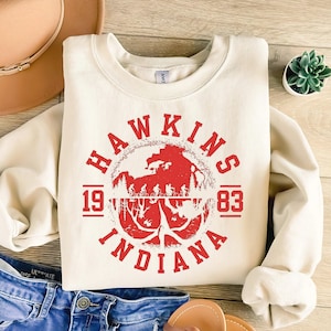May include: Cream-colored sweatshirt with a red graphic featuring the words "HAWKINS INDIANA 1983." The graphic includes a silhouette of a forest and figures. The sweatshirt is folded, with a hat and jeans visible in the background.