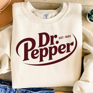 May include: Cream-colored sweatshirt featuring the Dr. Pepper logo in a deep burgundy color. The logo includes the brand name and the text "EST. 1885". The sweatshirt is a casual garment, suitable for everyday wear.