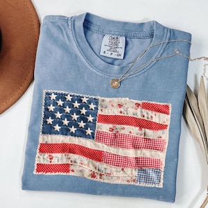May include: A pale blue Comfort Colors t-shirt with a patchwork American flag design. The flag features red floral, gingham, and solid fabrics for the stripes, with white stars on a blue background. A gold necklace with a pendant is draped across the shirt.