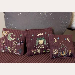 May include: Three small, square decorative pillows in varying sizes, each with a deep burgundy woven fabric. The pillows feature embroidered designs of moons, stars, and whimsical figures in shades of green, blue, and cream.