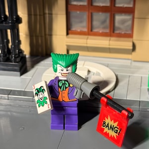 May include: A Lego figure of the Joker, with green hair, a purple suit, and a white face. He holds a playing card with a jester design and a toy gun with a red "BANG!" flag. The figure is set against a city backdrop.