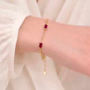 Peut inclure: Un bracelet doré orné de trois pierres précieuses rectangulaires rouge foncé. Le bracelet est délicat, avec un design en chaîne et un fermoir réglable. Les pierres sont espacées uniformément, créant un accessoire simple et élégant.