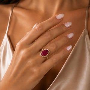 May include: A gold ring featuring a large, oval-shaped, deep red gemstone at its center. The central stone is surrounded by a halo of small, clear, sparkling stones set into the gold band, which continues down the sides of the ring setting.