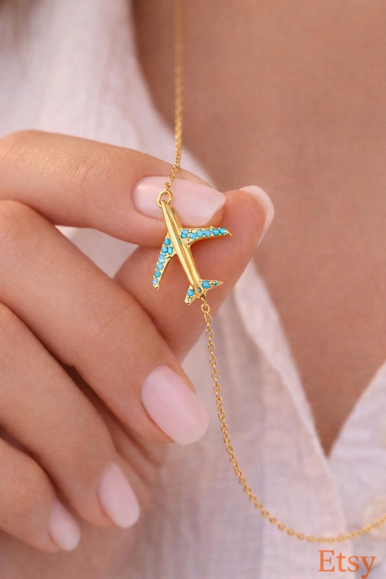 May include: A gold-colored airplane pendant necklace with turquoise-colored accents on the wings. The necklace is held up by a hand, showcasing the delicate chain and the airplane charm. The Etsy logo is visible in the bottom right corner.