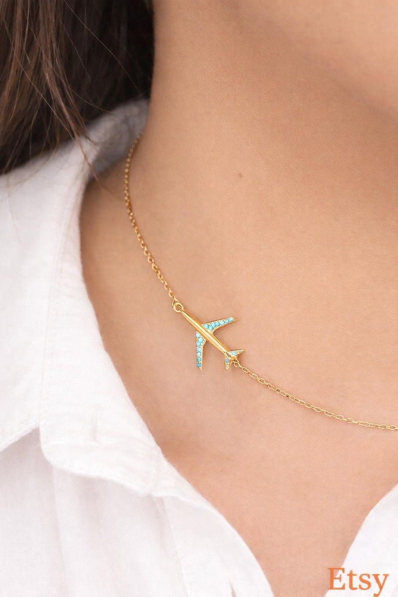 May include: A gold-colored airplane pendant necklace with turquoise accents. The necklace is worn on a person wearing a white shirt. The Etsy logo is visible in the bottom right corner.
