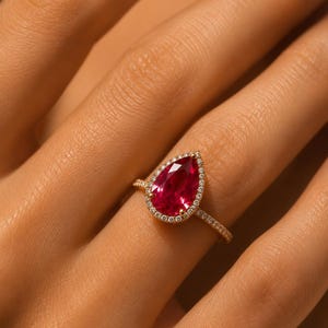 Op de afbeelding: Een gouden ring met een peervormige rode edelsteen als middelpunt, omringd door een halo van kleine witte diamanten. De dunne ringband is aan de bovenste helft ook bezet met kleine witte diamanten, wat zorgt voor een verfijnde uitstraling.