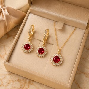 May include: A jewellery set consisting of a gold necklace and two matching hoop earrings. Each piece features a round, faceted red gemstone surrounded by a halo of small, clear white stones, all set in a polished gold-tone metal frame.