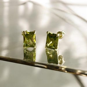 Può includere: Un paio di orecchini a lobo con gemme quadrate verde oliva. Le gemme sono incastonate in un metallo color oro. Gli orecchini sono esposti su una superficie riflettente. Gioielli.