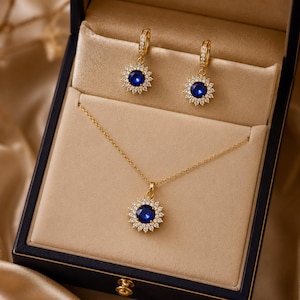 May include: A gold jewelry set consisting of a necklace and two hoop earrings, each featuring a central round deep blue stone surrounded by a halo of small clear white stones, presented in a dark blue box with a beige velvet interior lining.