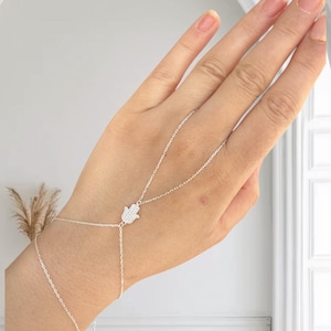 Puede incluir: Un brazalete de cadena de plata con un dije de mano de hamsa. El brazalete está diseñado para usarse en la muñeca y el dedo.