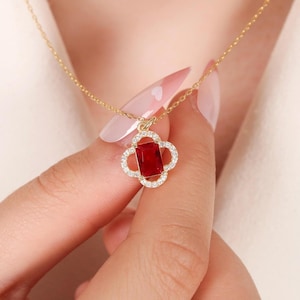 May include: A gold-colored necklace featuring a four-leaf clover pendant. The pendant has a rectangular, deep red gemstone at its center, surrounded by a halo of small, sparkling clear stones. The necklace is held by a person.