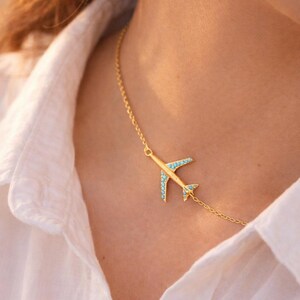 May include: Gold-colored airplane pendant necklace with turquoise-colored accents. The airplane design is on a delicate chain. The necklace is worn against a white shirt.