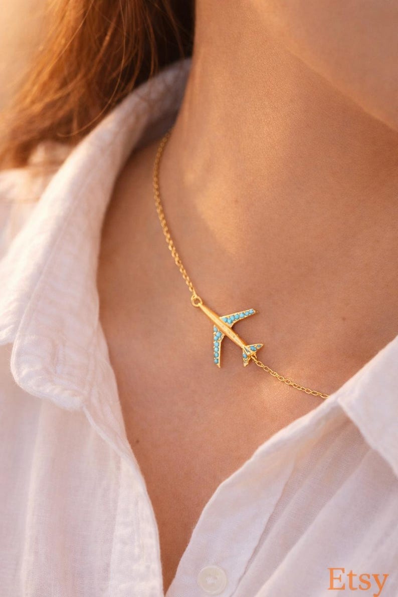 May include: Gold-colored airplane pendant necklace with turquoise-colored accents. The airplane design is on a delicate chain. The necklace is worn against a white shirt.