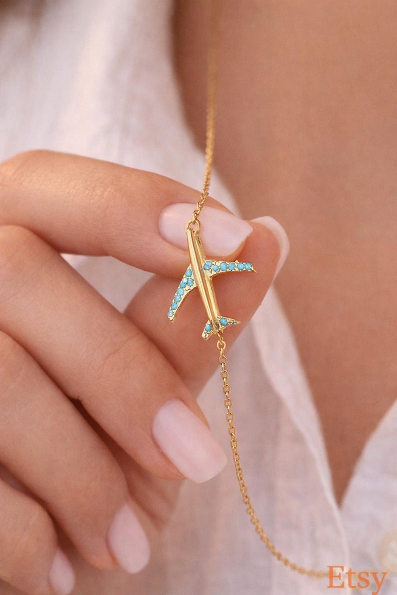 May include: A gold-colored airplane pendant necklace with turquoise-colored accents on the wings and tail. The necklace is on a delicate gold chain. The Etsy logo is visible in the bottom right corner.