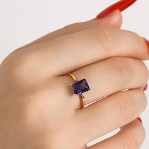 Pode incluir: Um anel dourado com uma pedra preciosa retangular roxa escura. O anel tem uma banda fina e simples, e a pedra preciosa é definida em um estilo minimalista. O anel é mostrado em uma mão com esmalte de unhas vermelho.