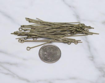 May include: A collection of brass-colored eye pins, each with a looped end, are scattered on a white marble surface. A U.S. dime is included for size reference. These pins are likely used for jewelry making or crafting.