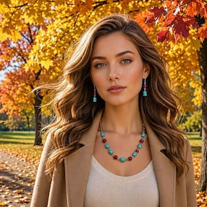 May include: A necklace and matching drop earrings featuring alternating turquoise-coloured beads and dark red spherical beads on a metallic chain. The jewellery is worn with a white top and a tan coat against a backdrop of autumn trees.